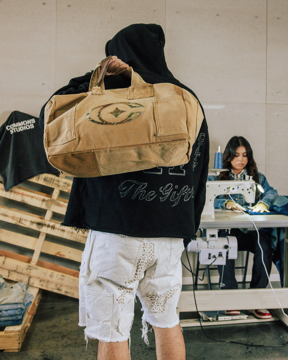 Person carrying a tan canvas duffel bag with handles, patch pockets, and a faded oval logo in a sewing workshop.