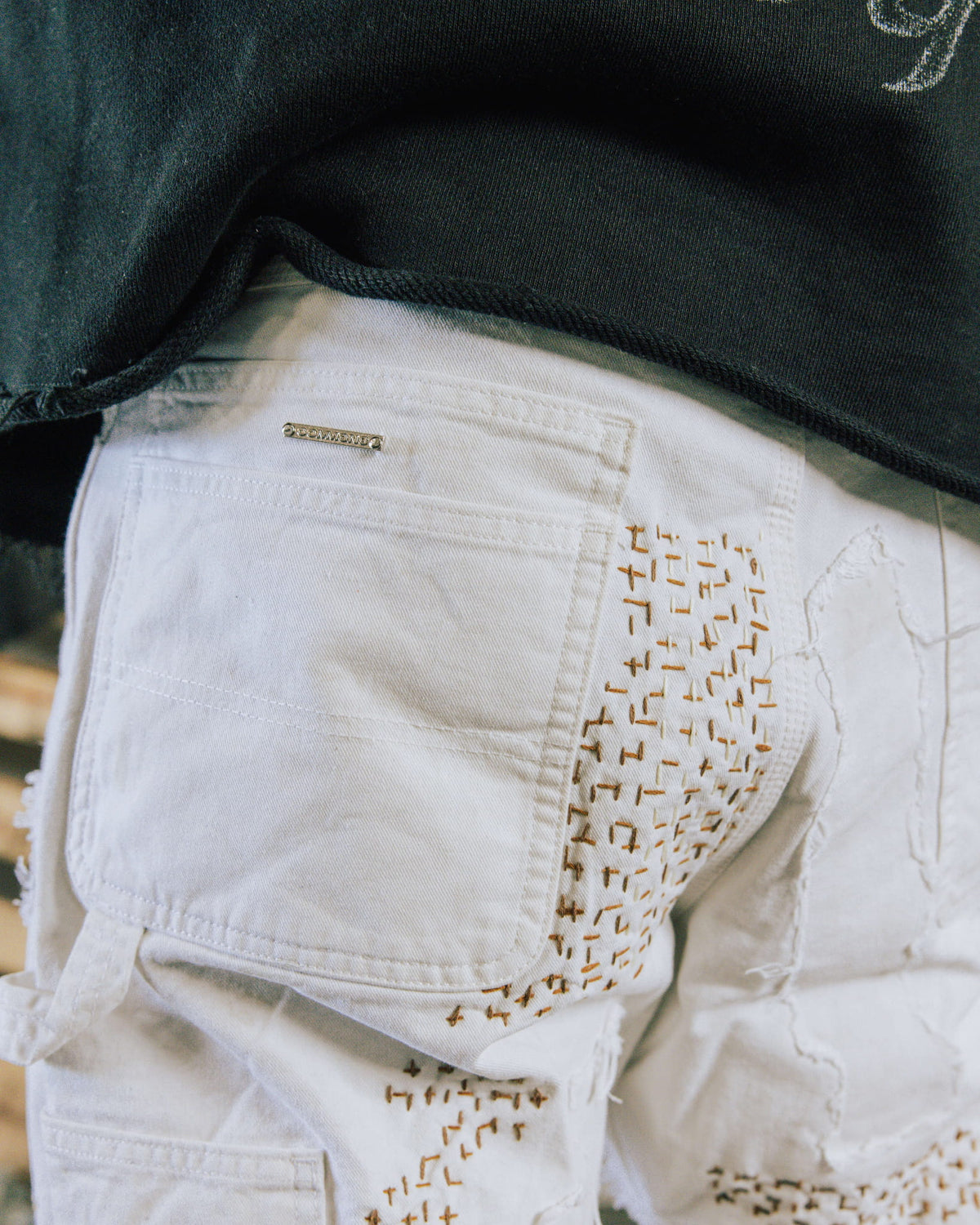 Close-up of white denim pants with a back pocket, brown decorative stitching, and frayed patches beneath a black top.