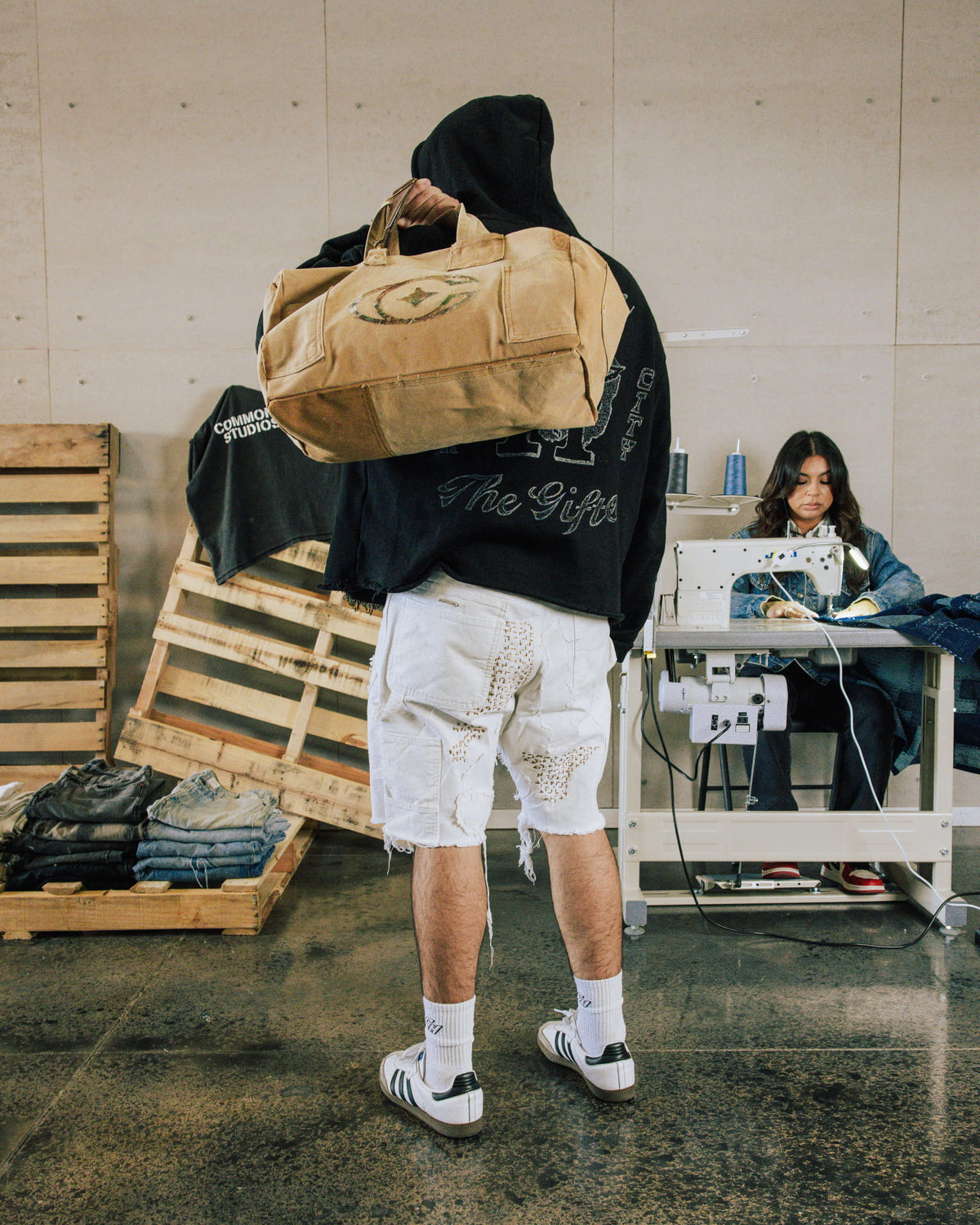 Person in a black hoodie and distressed white denim shorts carrying a tan canvas duffel bag in a sewing workshop.