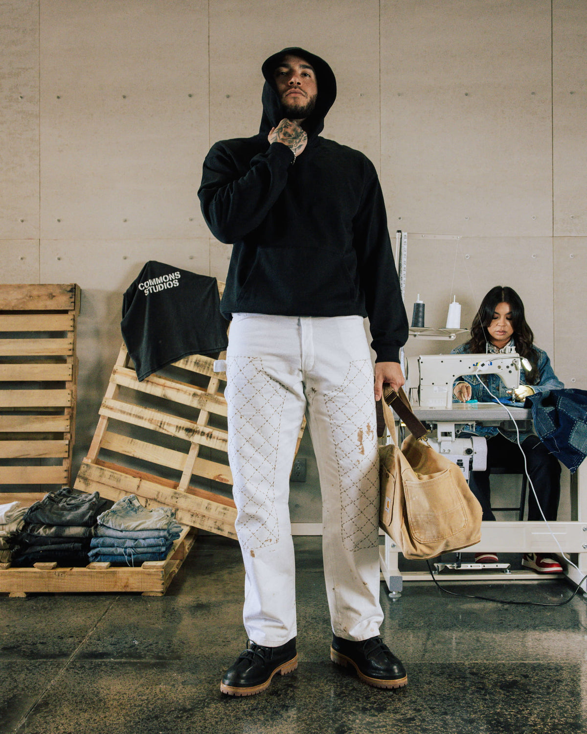 Man in a black hoodie, white patterned pants and black boots holding a tan bag in a sewing workshop.