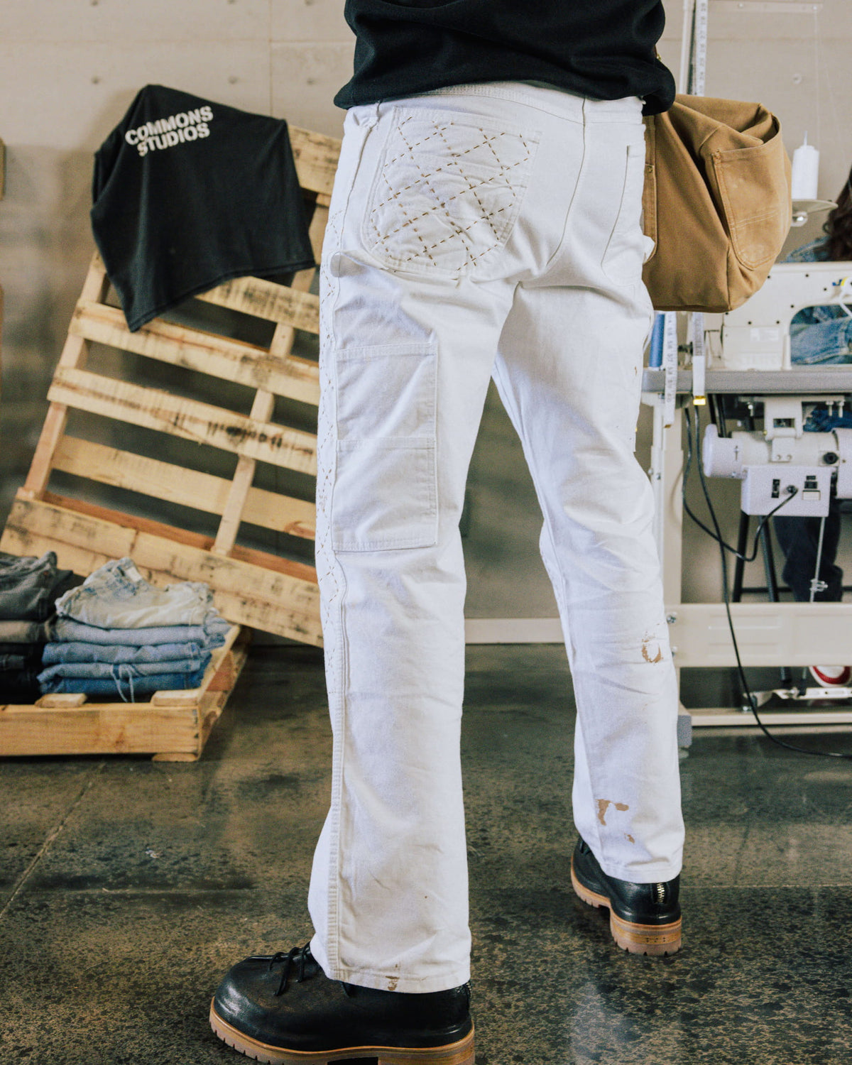 Person wearing white work pants with stitched back pockets and tan marks, black boots, and a tan bag in a sewing workshop.