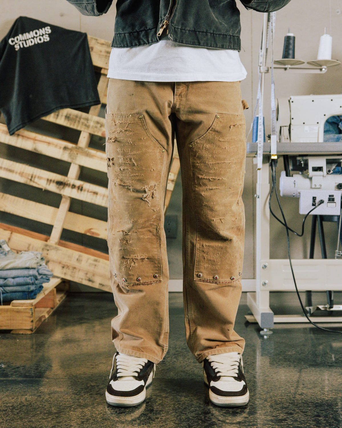 Person wearing distressed tan work pants with riveted knee panels and black-and-white sneakers in a sewing workshop.