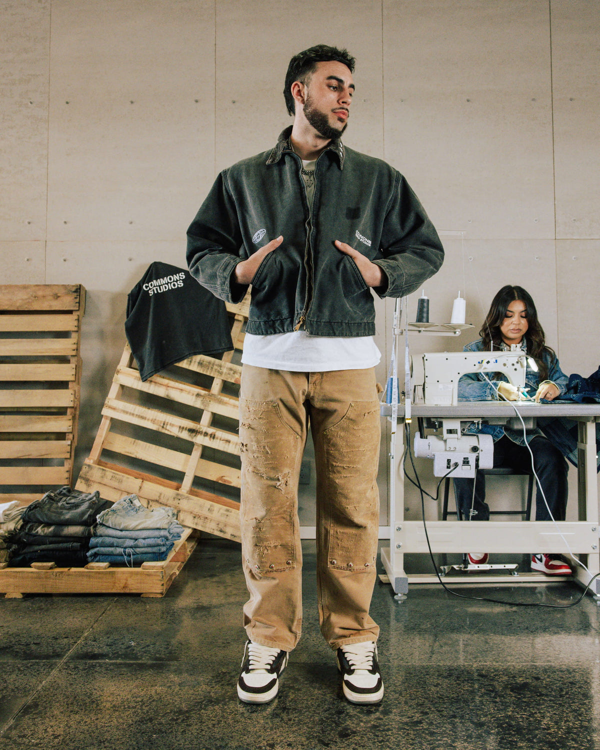 Man wearing a faded black denim jacket, distressed tan pants and black-and-white sneakers in a sewing workshop.