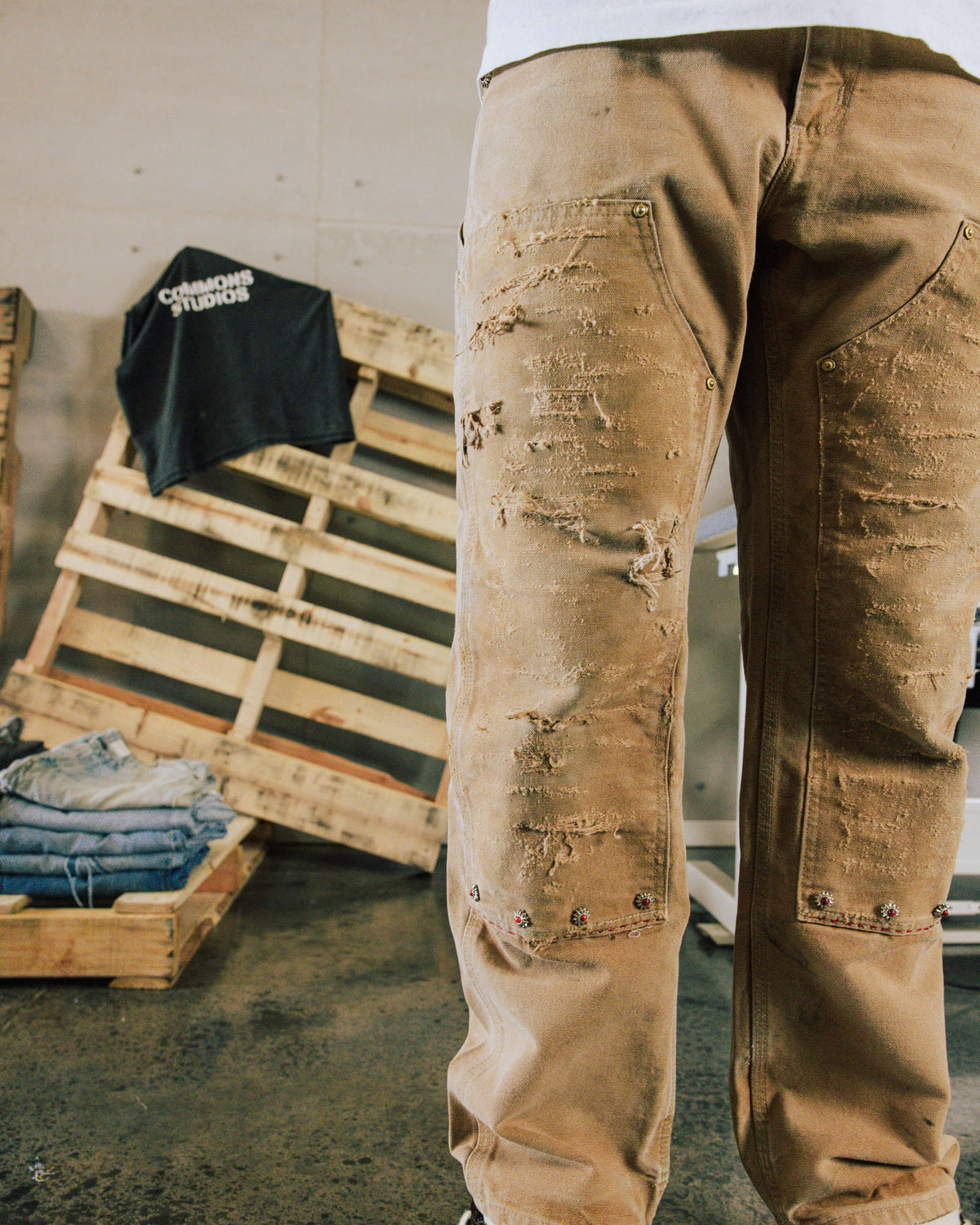 Tan distressed pants with riveted front panels and decorative studs, worn beside wooden pallets and folded jeans.