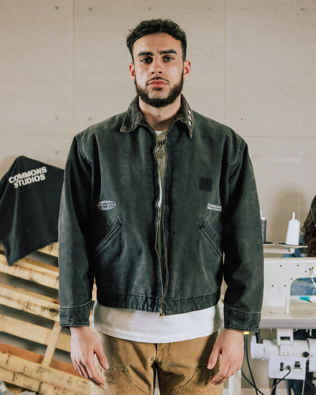 Man wearing a faded charcoal zip-front jacket with white logos and slanted pockets in a sewing studio.