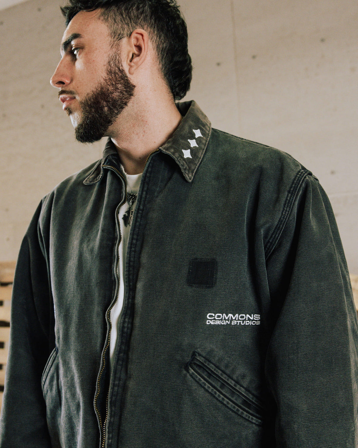 Man wearing a washed charcoal zip-front jacket with white collar motifs, embroidered branding, and angled pockets.