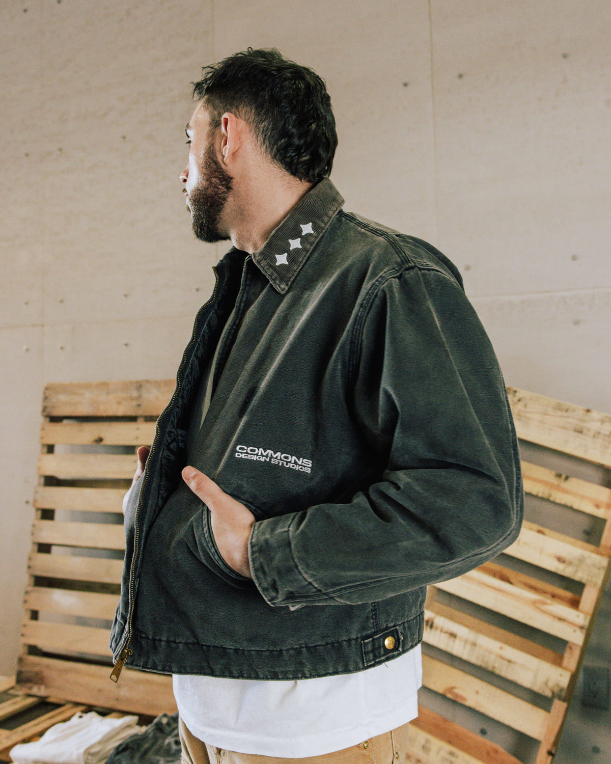 Man wearing a washed charcoal zip jacket with white chest lettering and three collar stars, standing beside wooden pallets.