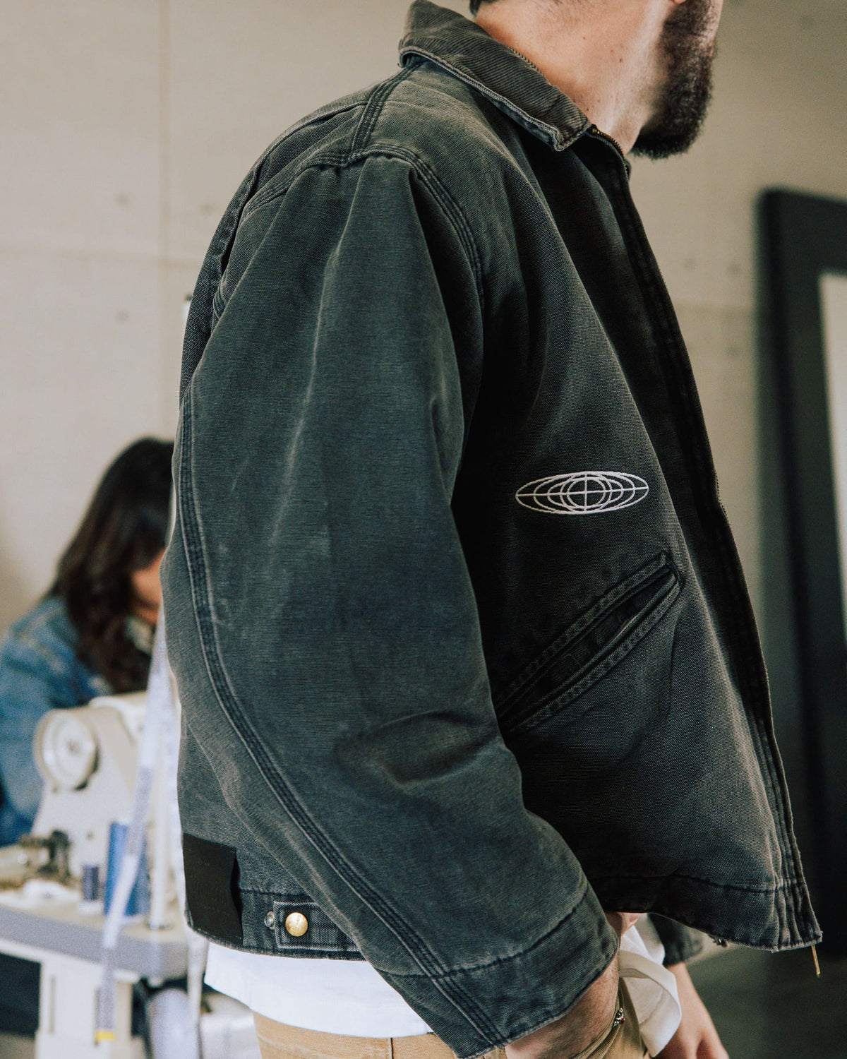 Person wearing a faded charcoal canvas jacket with a white embroidered emblem and angled pocket in a sewing workshop.