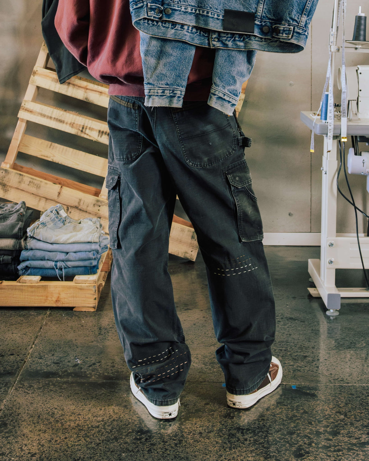 Person wearing faded black cargo jeans with contrast stitch details and a burgundy sweatshirt in a sewing workshop.