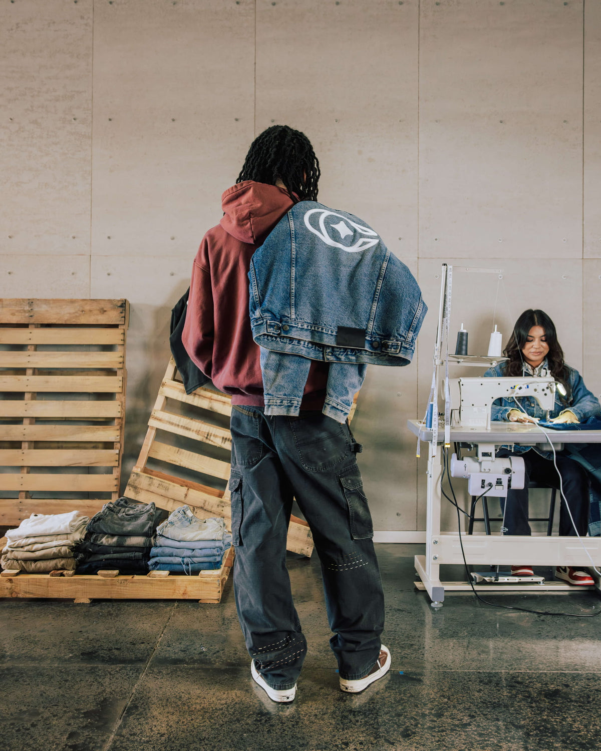 Person in a rust hoodie and dark cargo jeans carries a blue denim jacket with a white graphic in a sewing workshop.