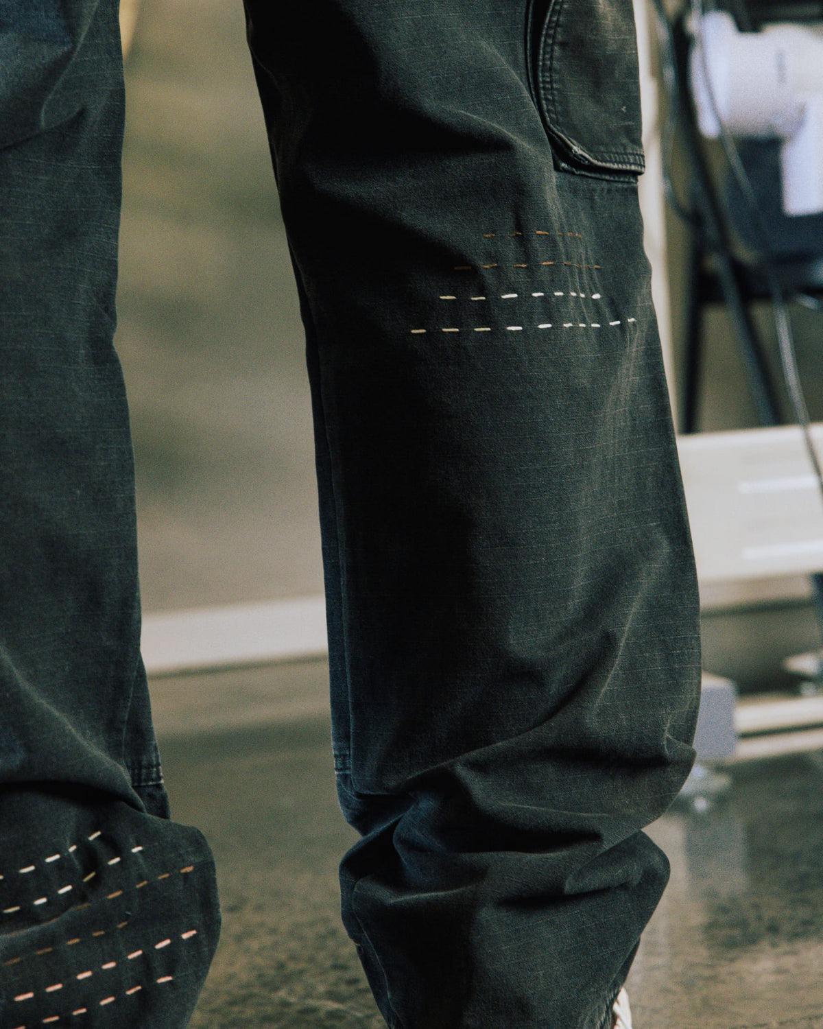 Close-up of dark denim pants with a patch pocket and rows of white and tan decorative stitching on the legs.