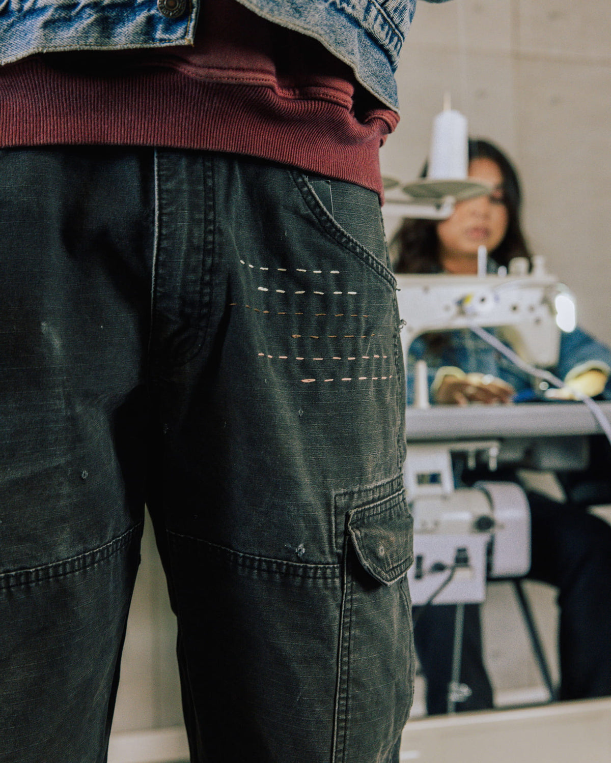 Dark faded cargo pants with contrast stitch details and a flap pocket, worn near a sewing machine.