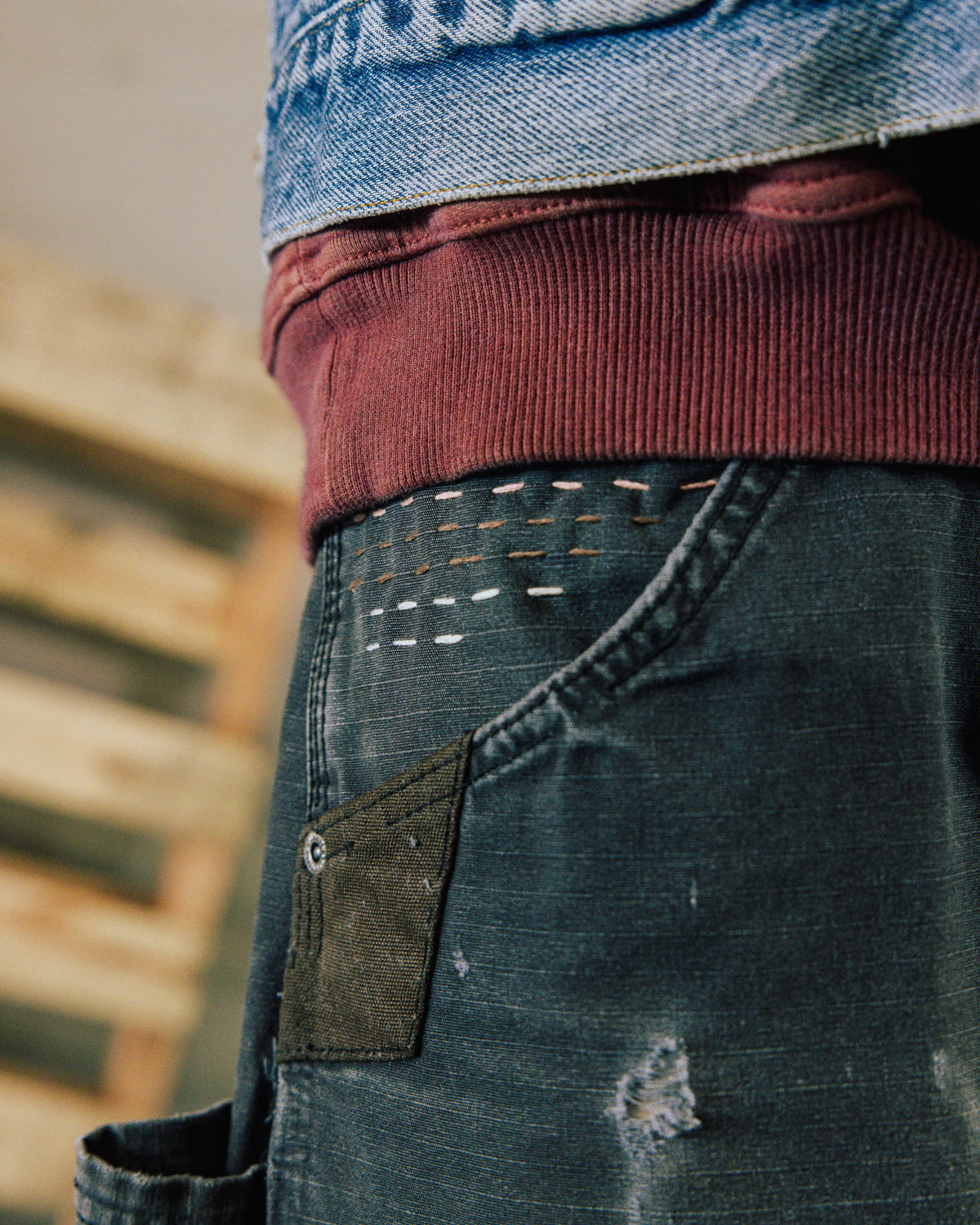 Close-up of dark blue distressed denim pants with contrast stitching and a riveted patch beneath a burgundy ribbed hem.