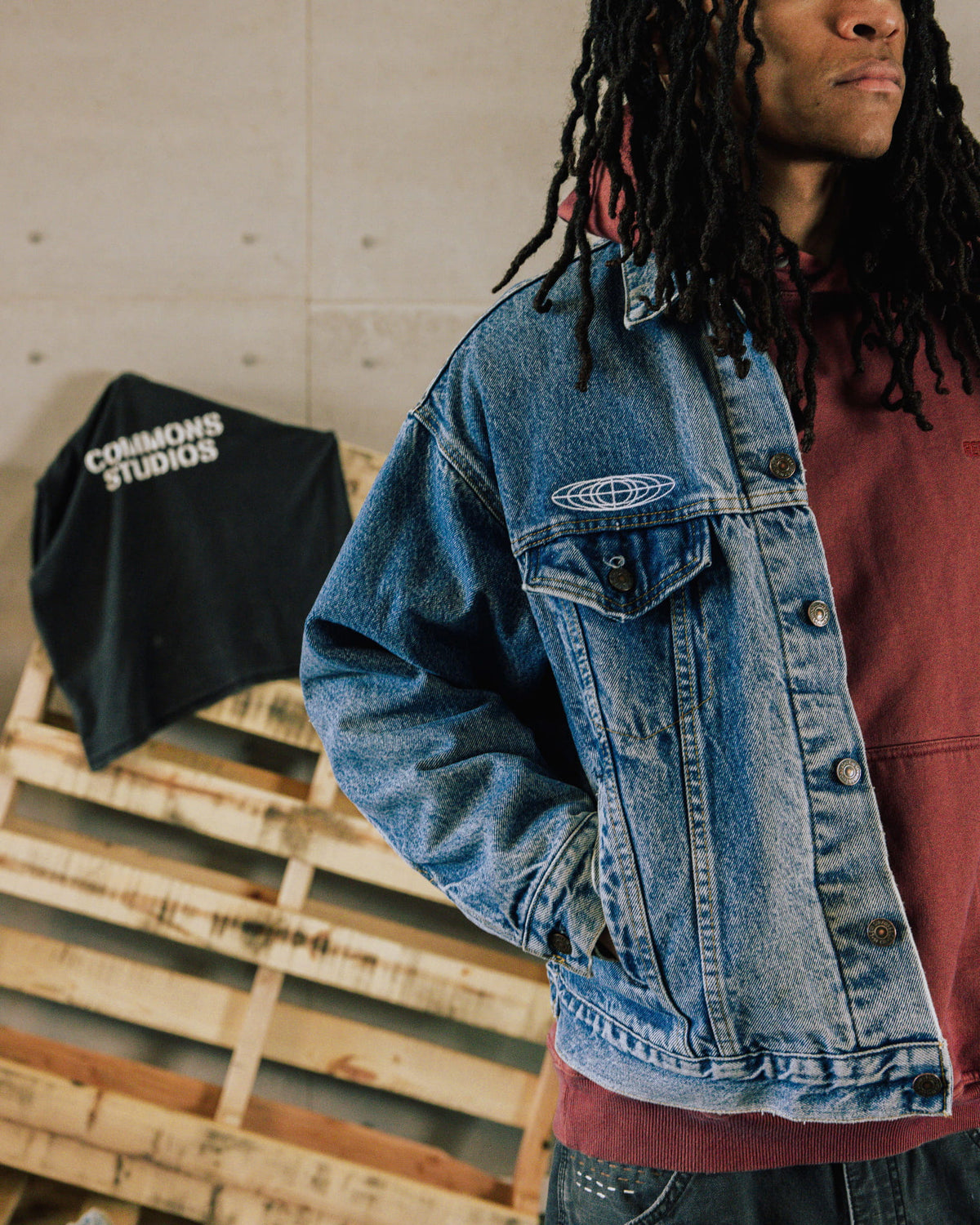 Person wearing a blue denim jacket with white chest embroidery over a burgundy hoodie beside wooden pallets.