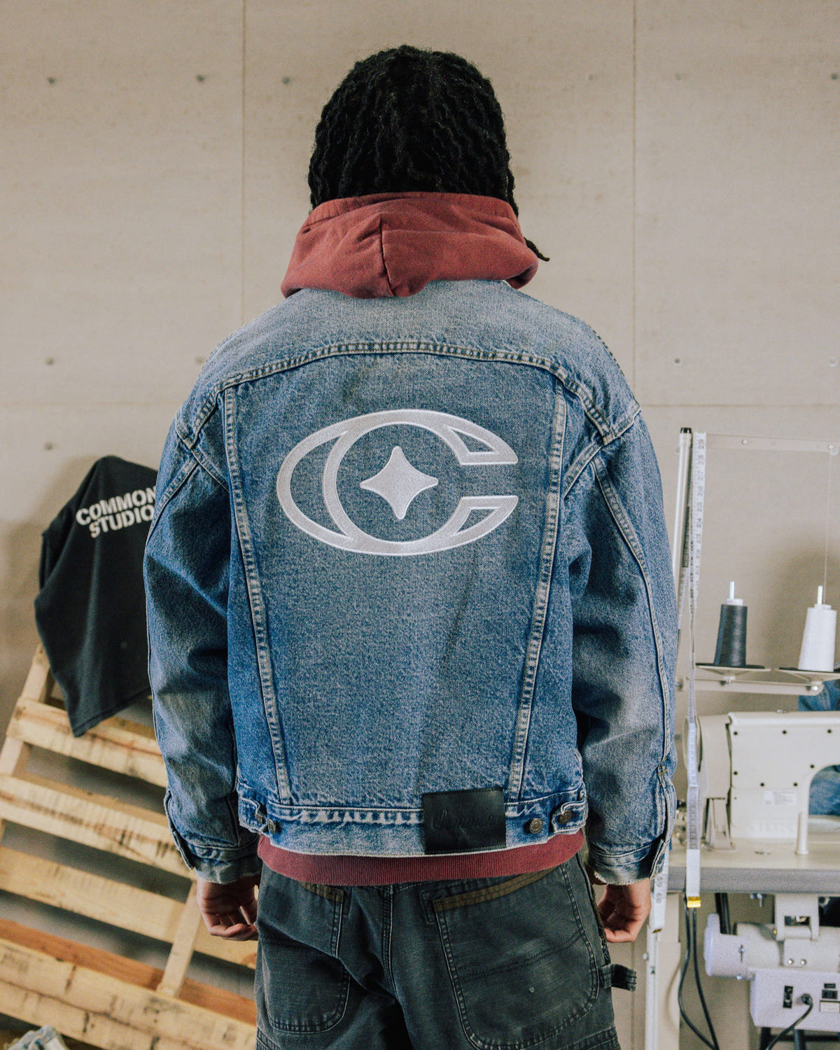 Person facing away in a blue denim jacket with a large white back emblem over a rust hoodie in a sewing studio.