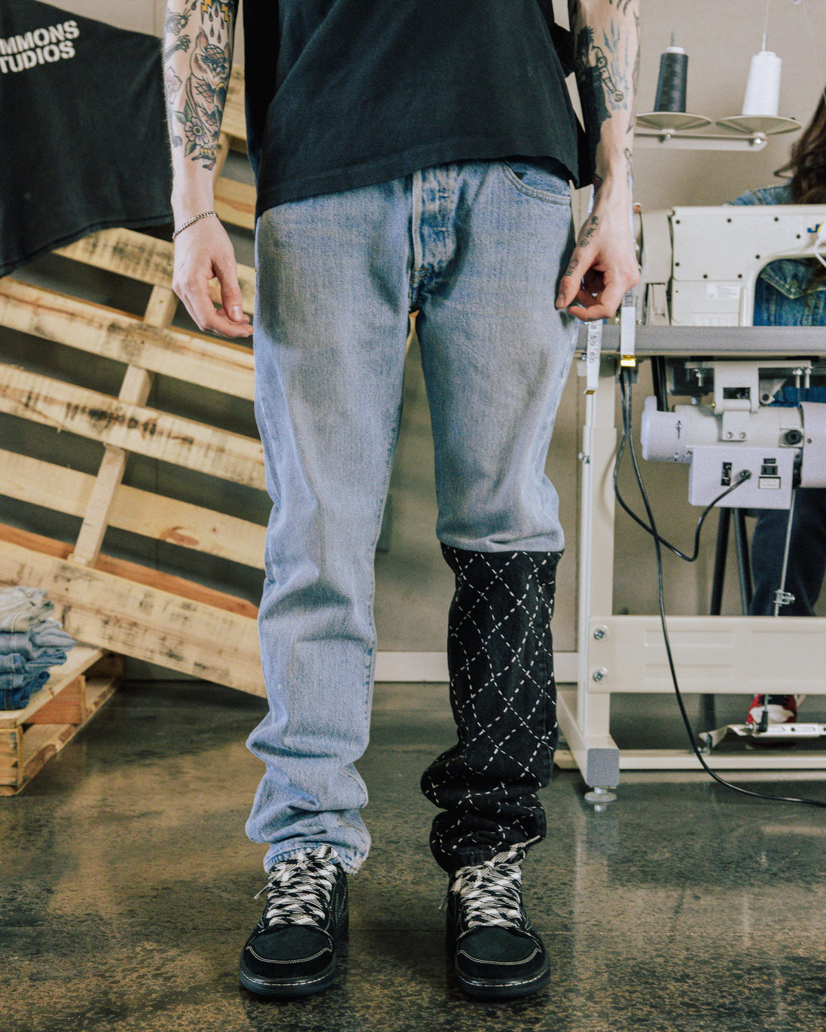 Model wears light blue denim jeans with a black, white-stitched lower leg panel beside a sewing machine.