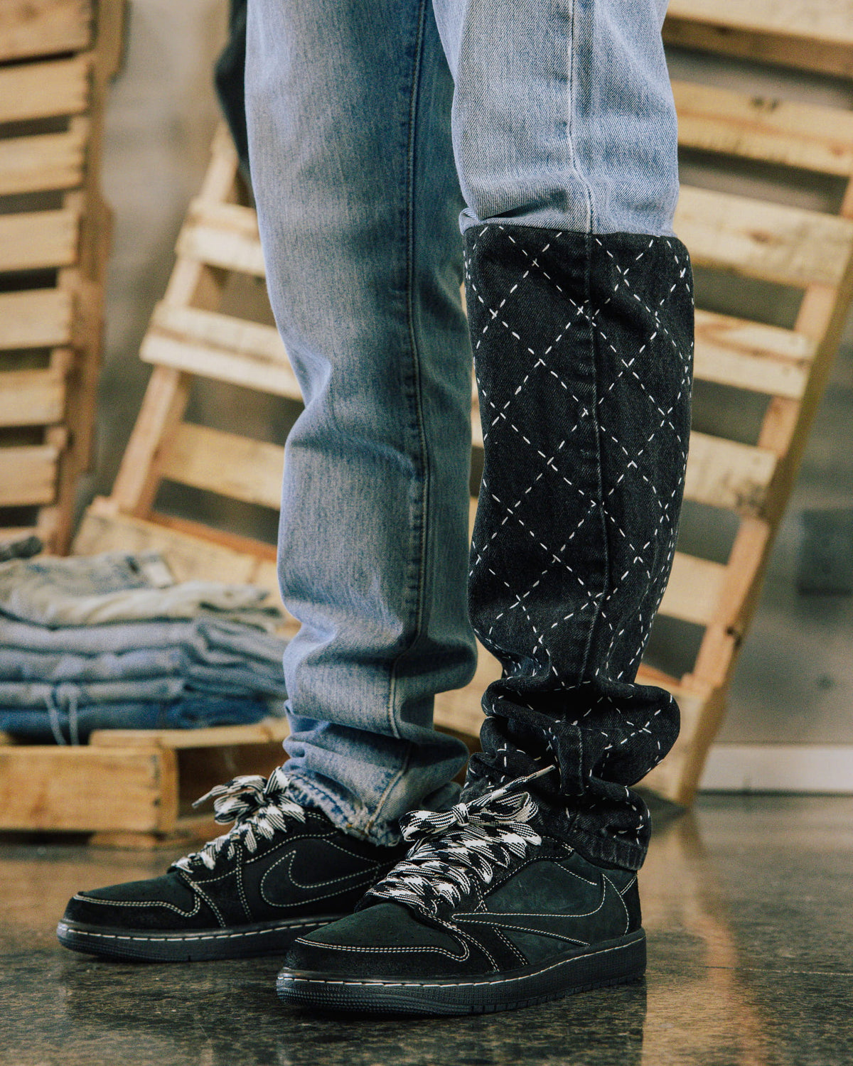 Light blue jeans with a dark denim leg panel and white diamond stitching, worn with black sneakers near wooden crates.