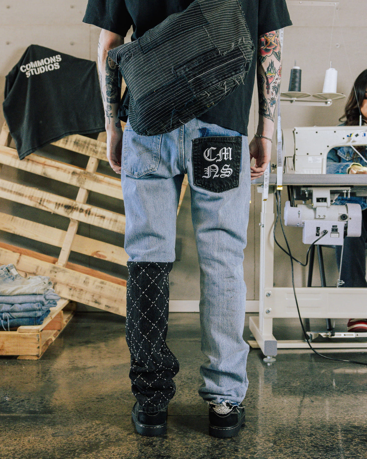 Person wearing light blue patchwork jeans with a black stitched leg panel and a black striped bag in a sewing studio.