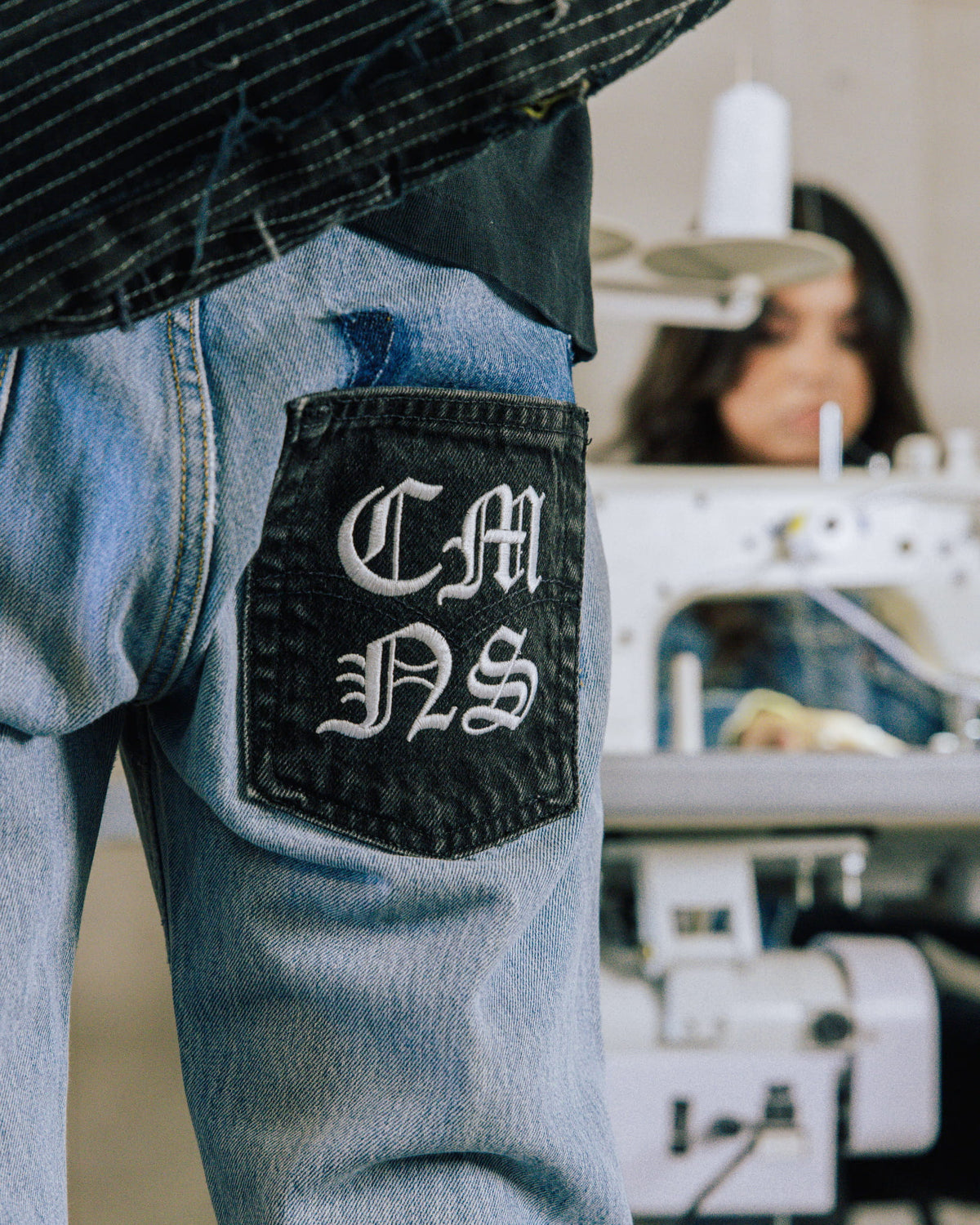 Light blue denim jeans with a black back pocket and white Gothic embroidery in a sewing workshop.