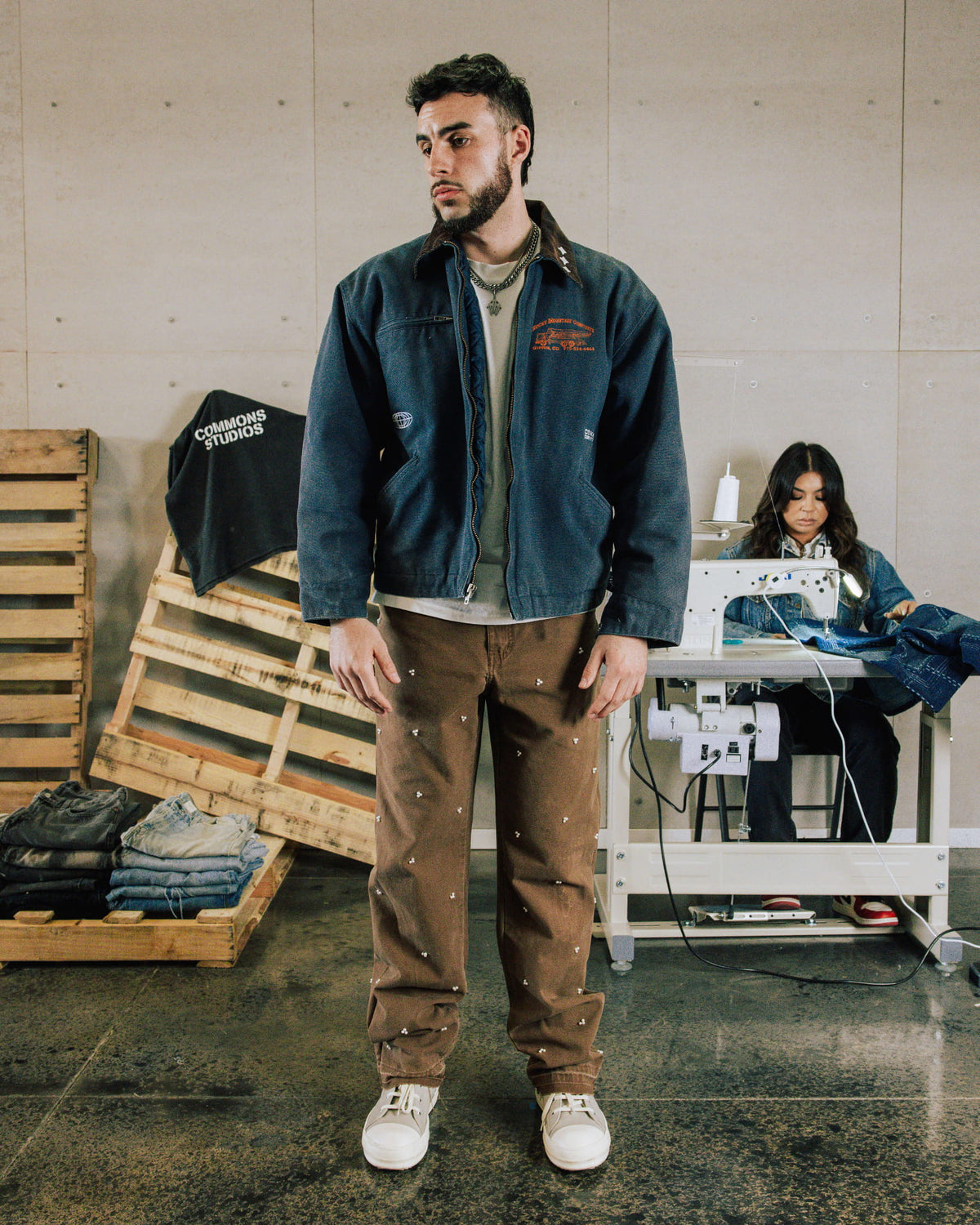 Man wearing a dark blue zip-up denim jacket and brown patterned trousers in a sewing workshop.
