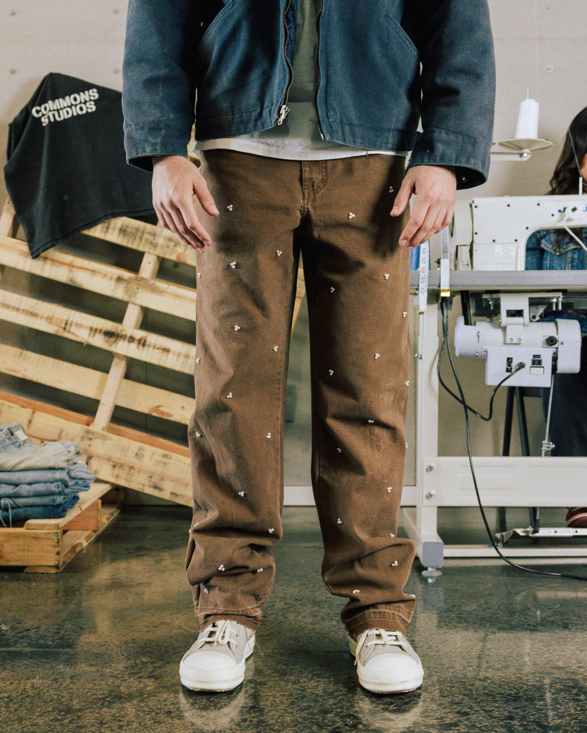 Person wearing brown pants with small white motifs, a dark blue zip jacket and white sneakers in a sewing studio.