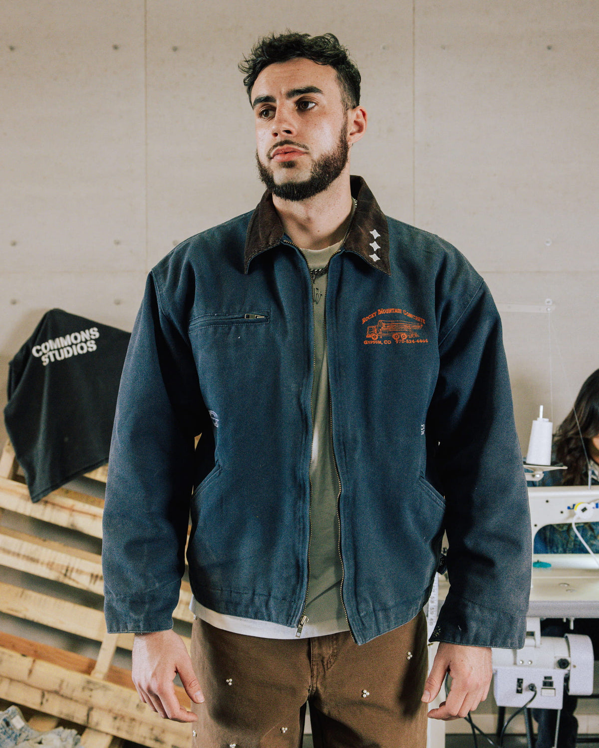 Man wearing a navy zip-front work jacket with a brown collar and orange chest embroidery in a sewing workshop.