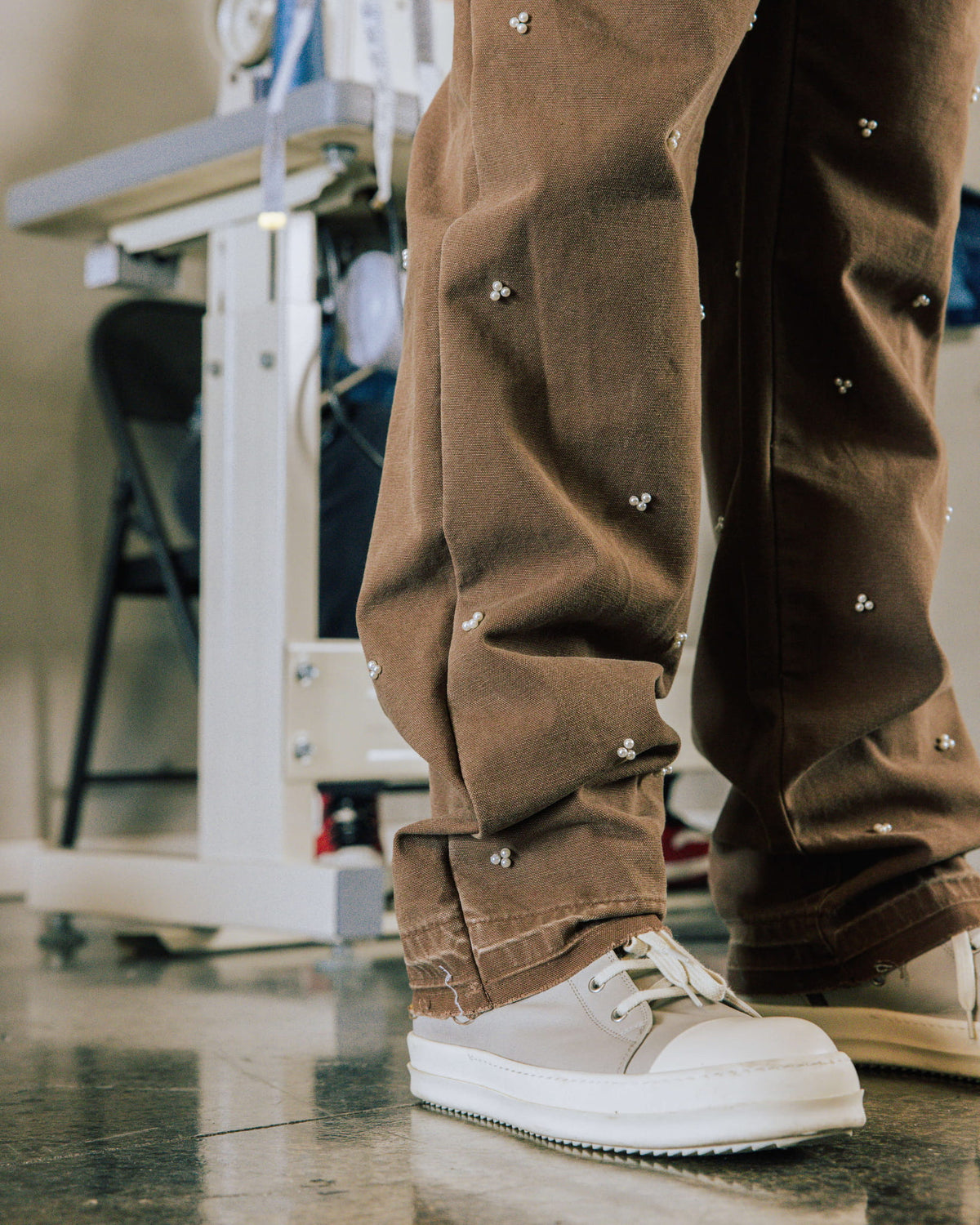 Brown trousers with small pearl-like bead clusters and wide hems, worn with chunky white lace-up shoes indoors.