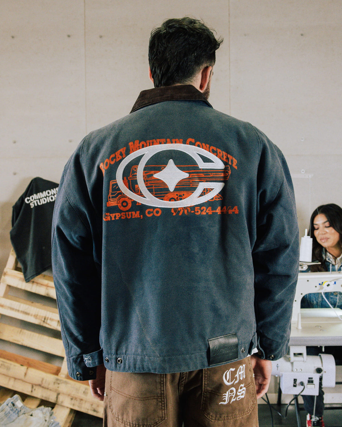 Man seen from behind wearing a dark gray jacket with a brown corduroy collar and orange and white back graphics in a workshop.