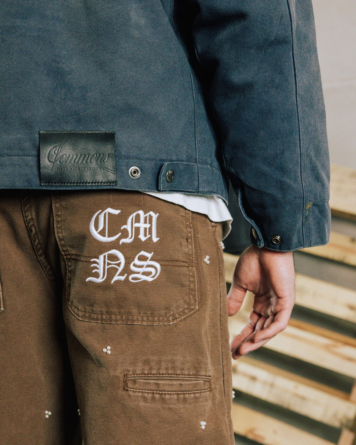 Person wearing a blue woven jacket with a black logo patch and brown pants with white pocket embroidery and bead accents.