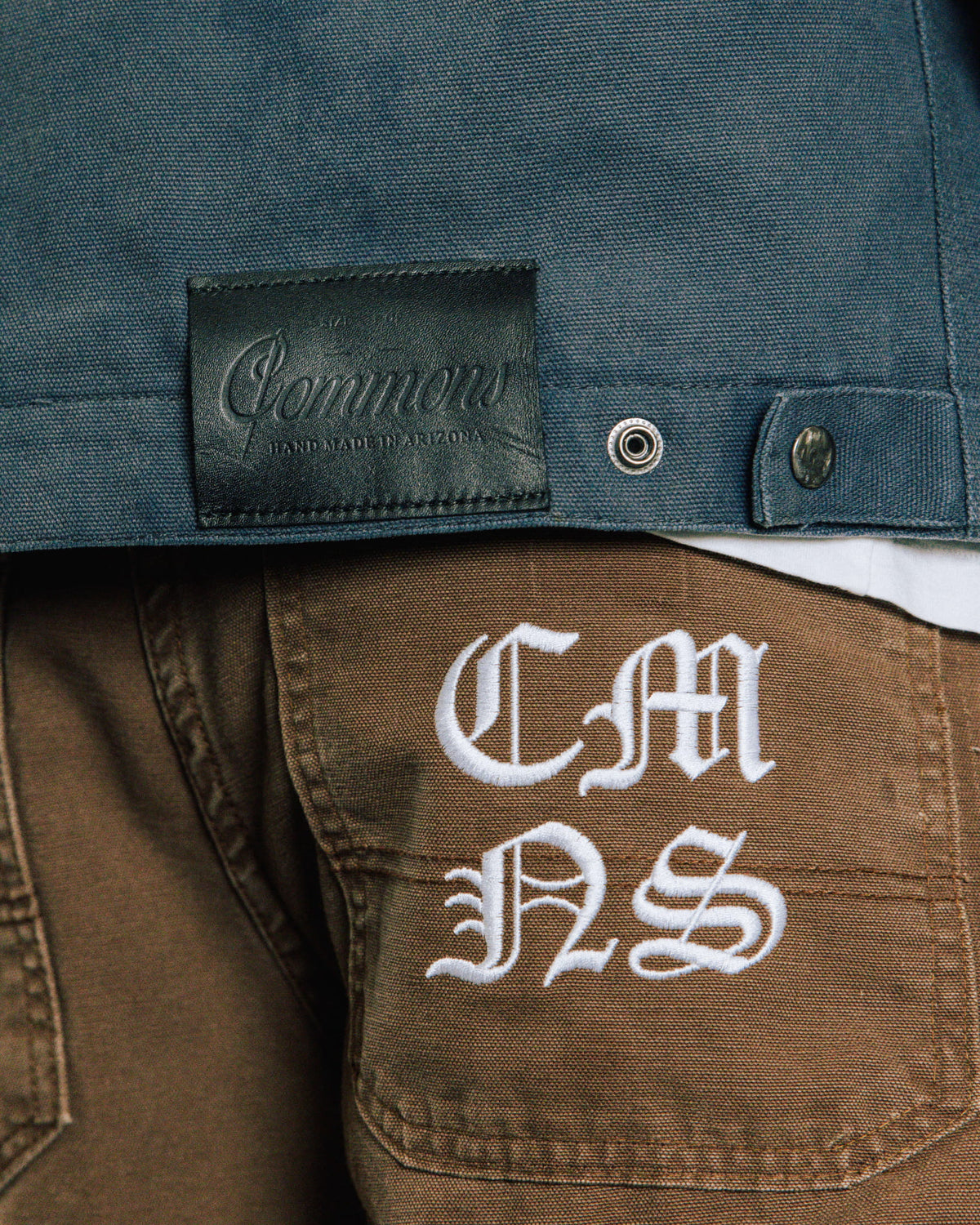 Close-up of a blue canvas jacket with a black logo patch above brown pants with white embroidered letters on a back pocket.