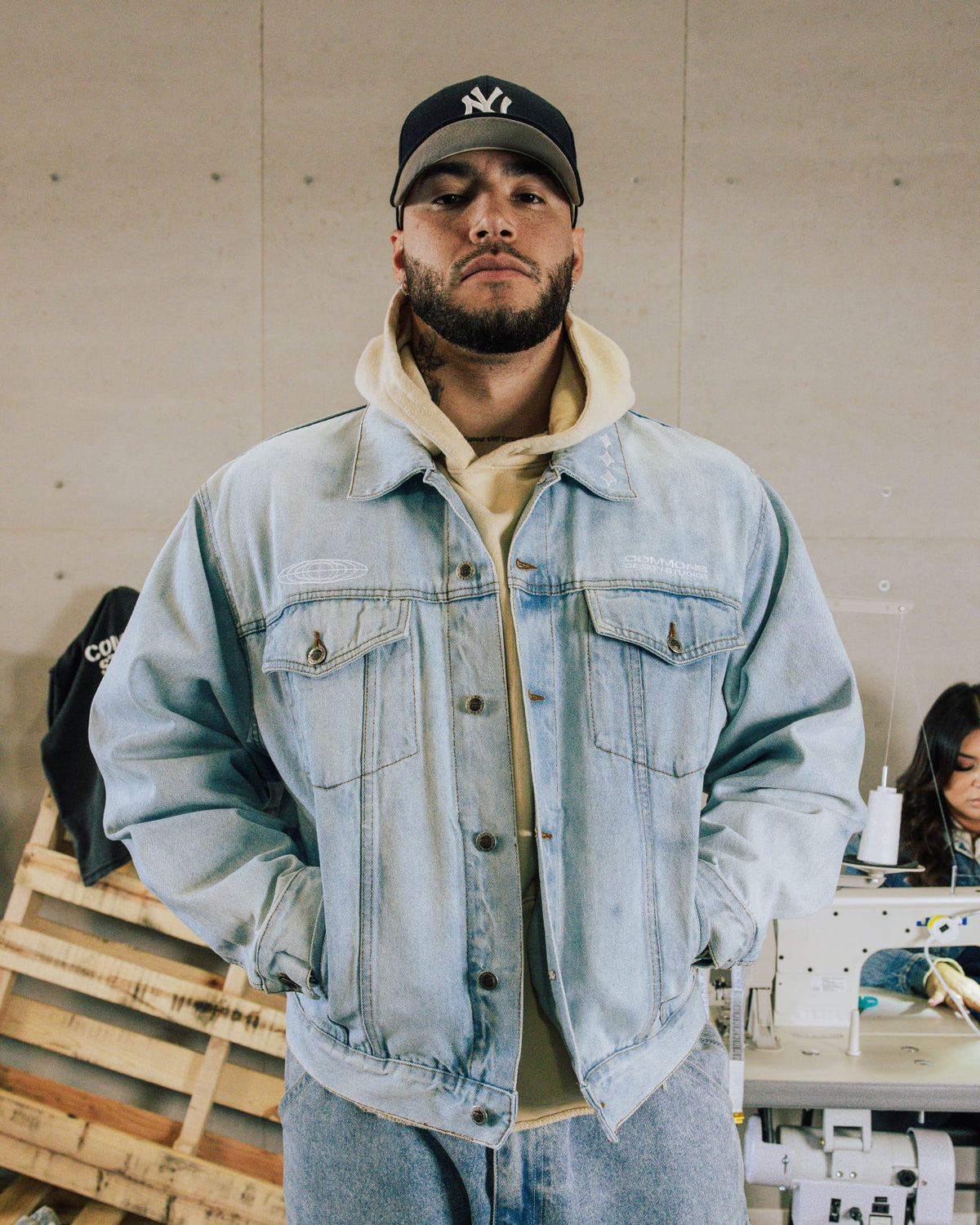Man wearing a light blue denim jacket with flap pockets over a cream hoodie and black cap in a sewing workshop.