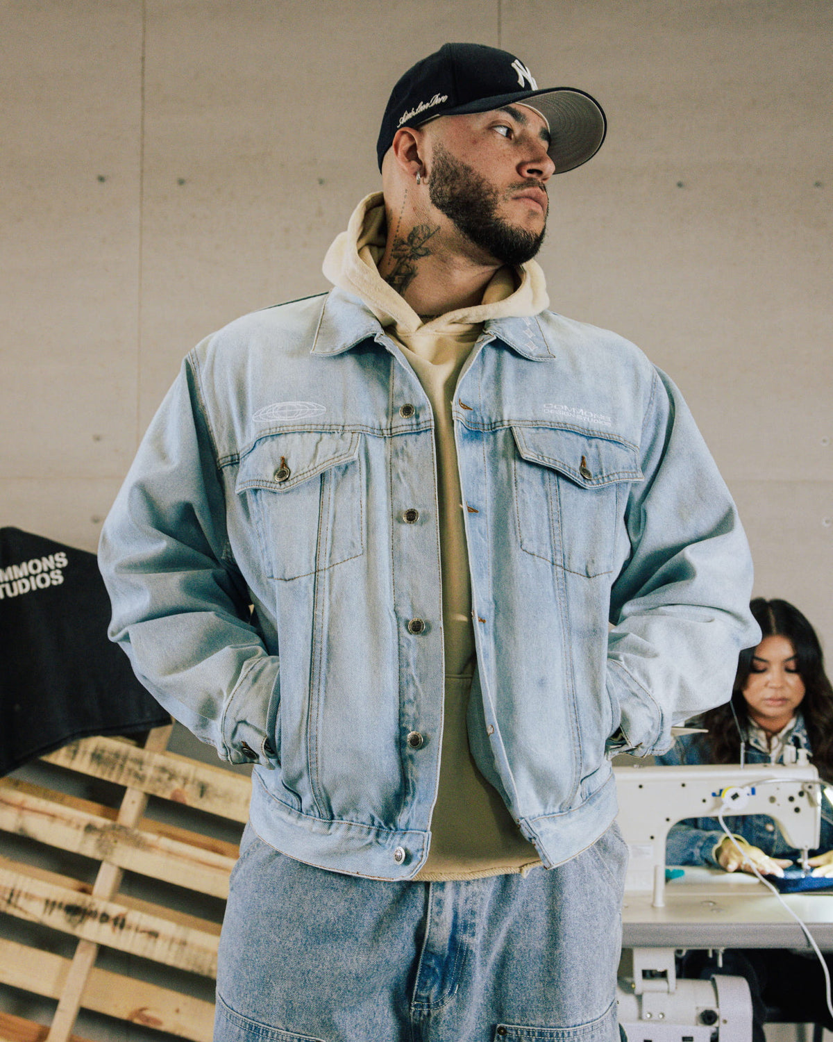 Man wearing a light blue denim jacket with flap pockets over a beige hoodie and black cap in a sewing workshop.
