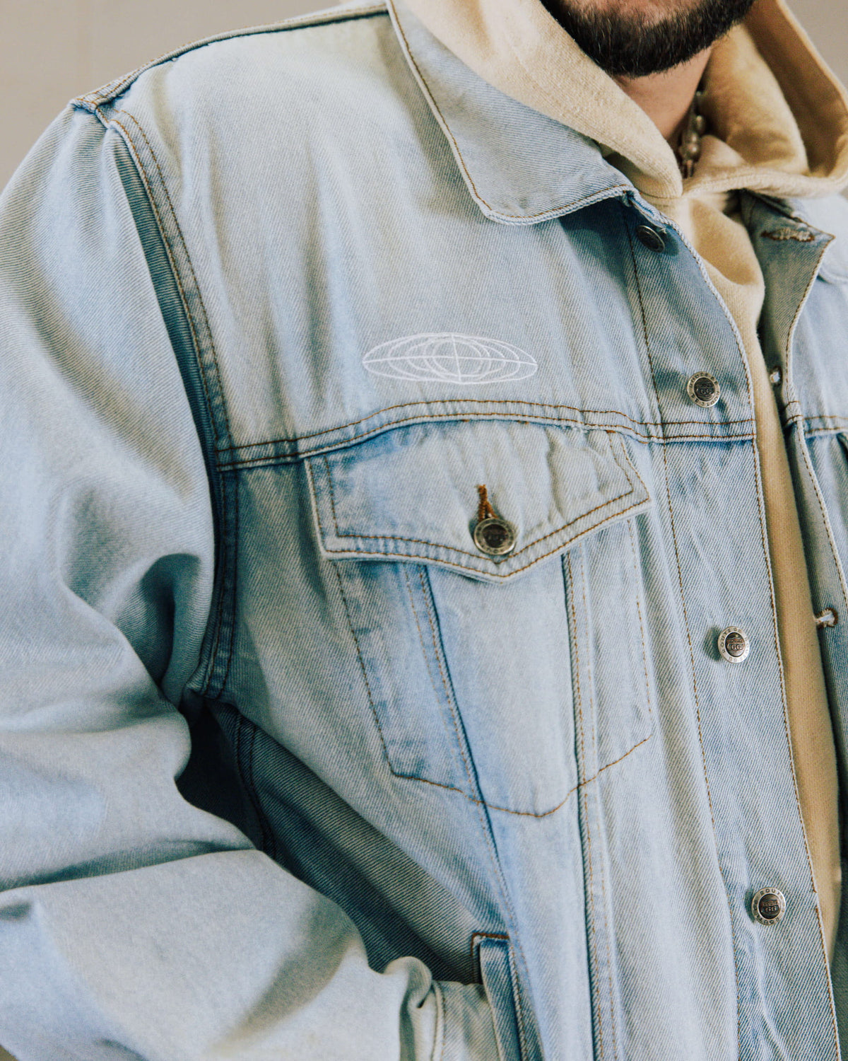Light blue denim jacket with white globe embroidery, metal buttons and a flap chest pocket, worn over a beige hoodie.