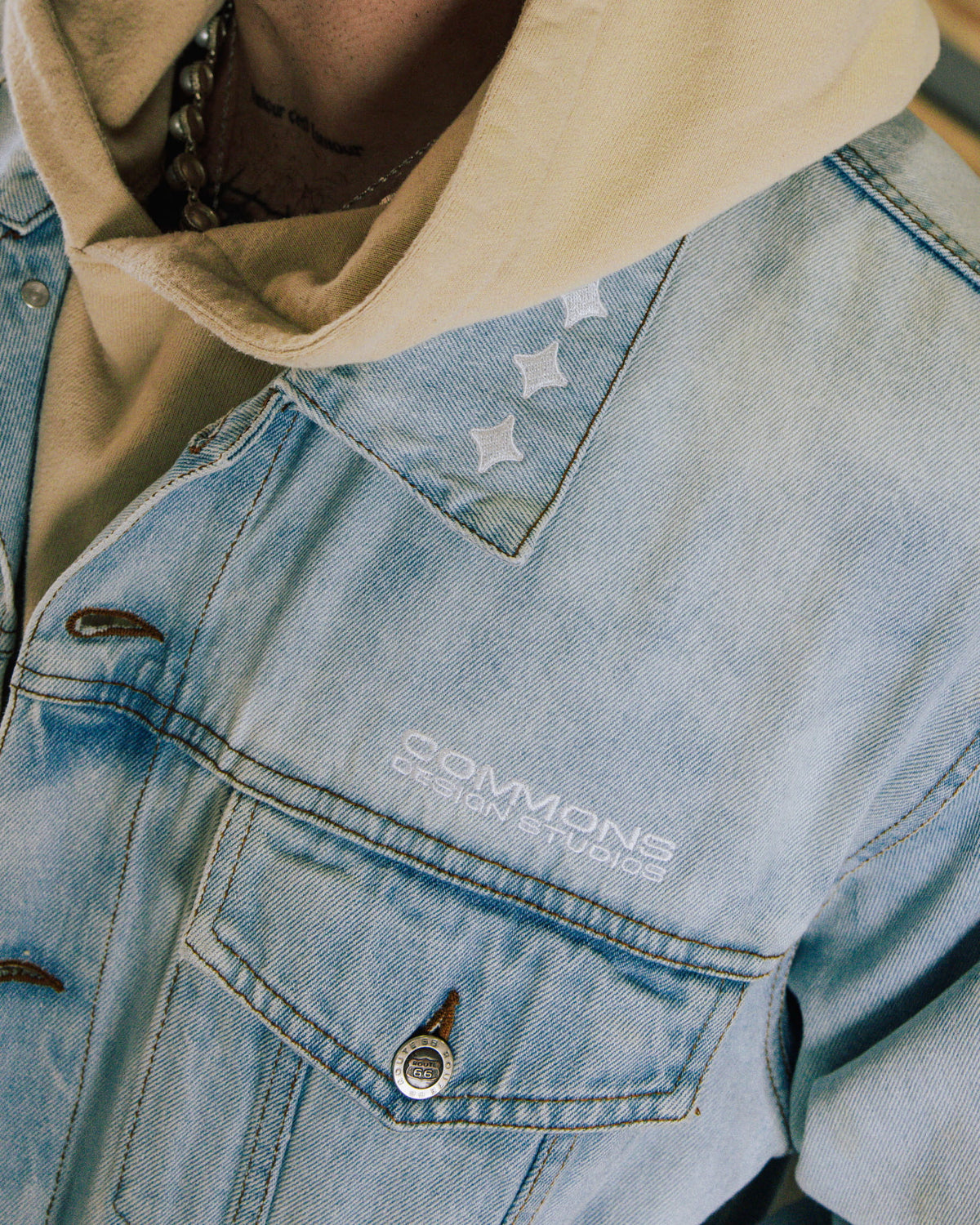Light blue denim jacket with white embroidered details and a buttoned chest pocket, worn over a beige hoodie.