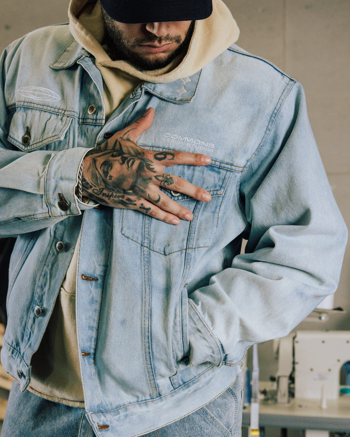 Man wearing a light blue denim jacket with white embroidery and flap pockets over a beige hoodie, with a black cap.