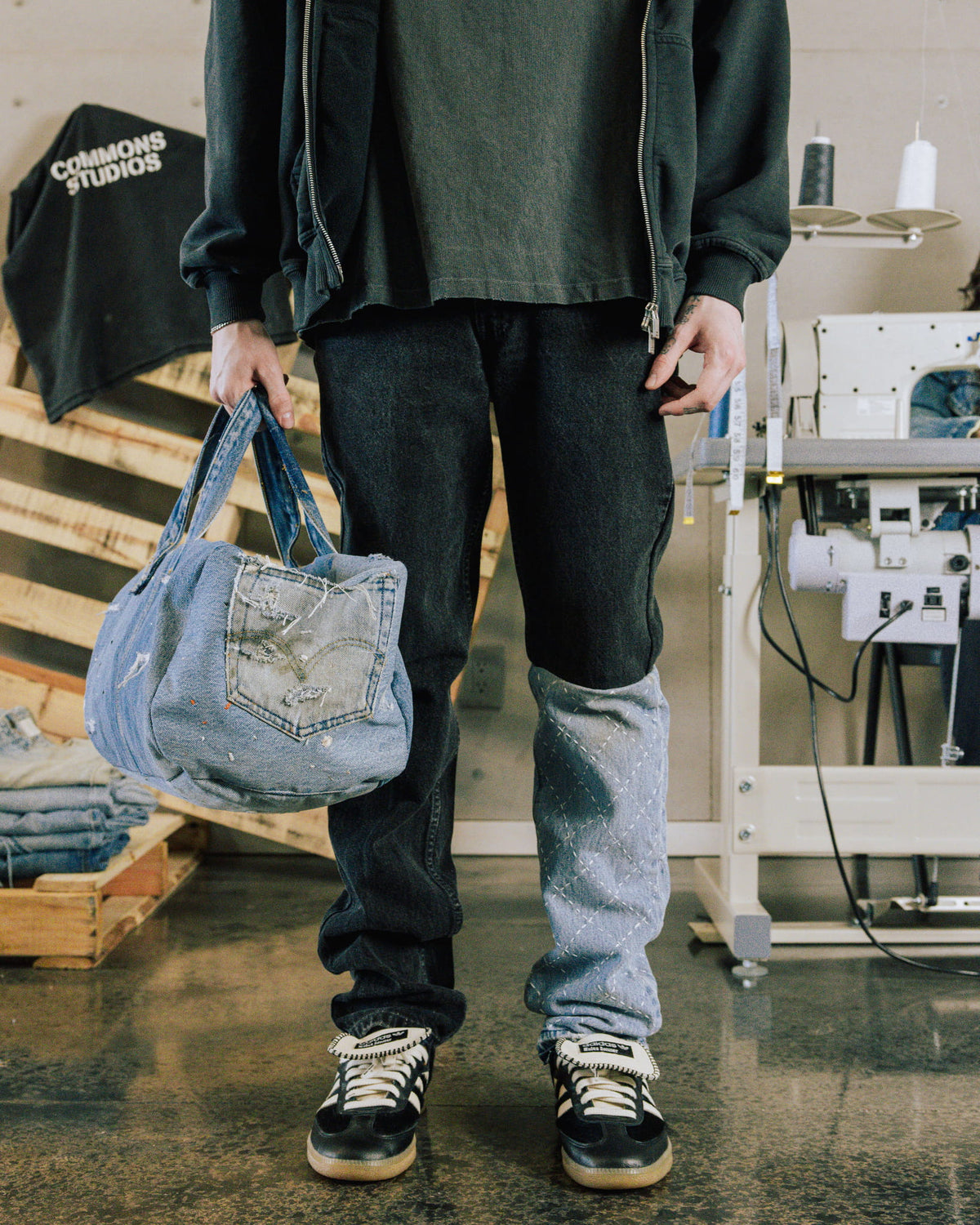 Person wearing dark jeans with a light blue stitched leg panel, holding a distressed denim bag in a sewing studio.