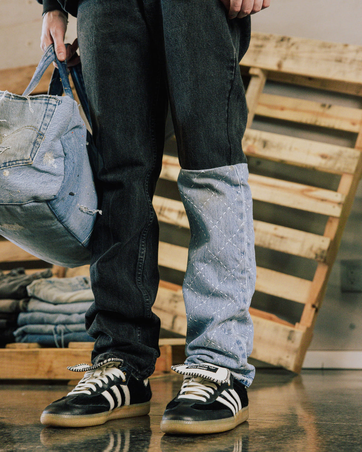 Person wears dark jeans with a light blue stitched leg panel and black sneakers, holding a distressed denim bag.