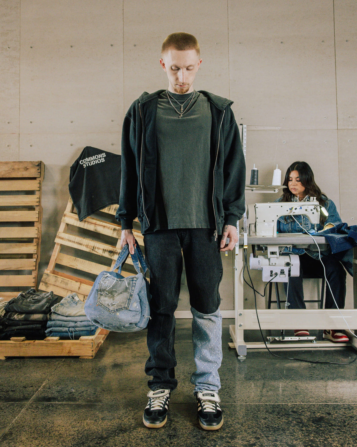 Model in a black hoodie and two-tone patchwork jeans holding a distressed blue denim bag in a sewing workshop.