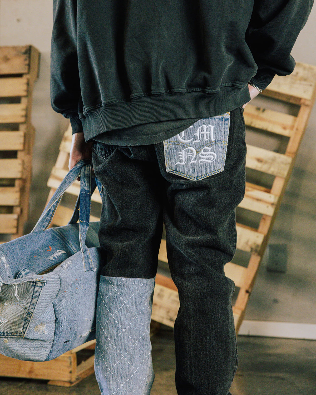 Person in a black sweatshirt and dark patchwork jeans with a lettered back pocket, holding a distressed denim bag.