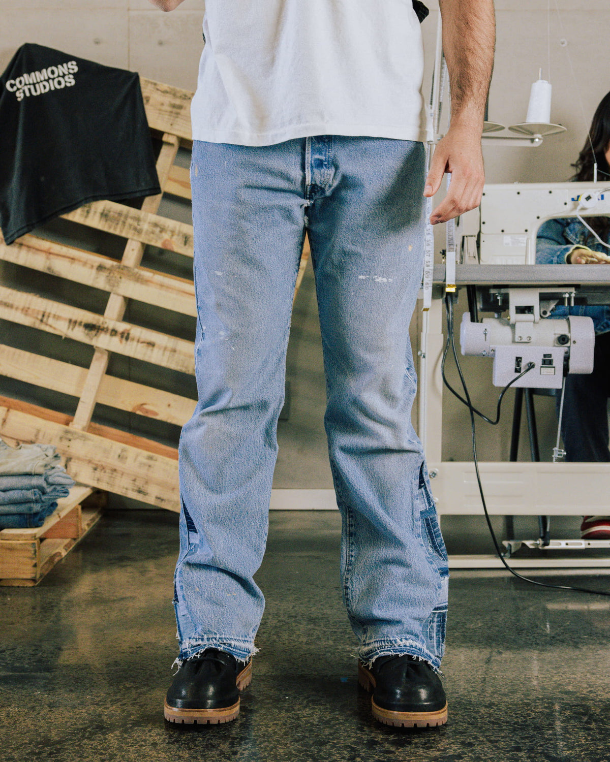 Person wearing light blue denim jeans with patchwork and frayed hems, a white T-shirt and black shoes in a sewing studio.