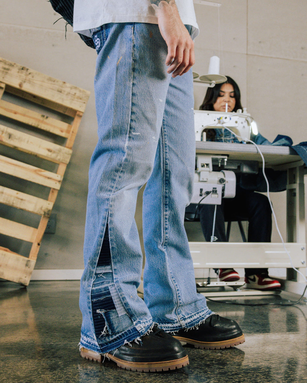 Light blue denim jeans with frayed hems and dark inset side slits, worn with black shoes in a sewing workshop.