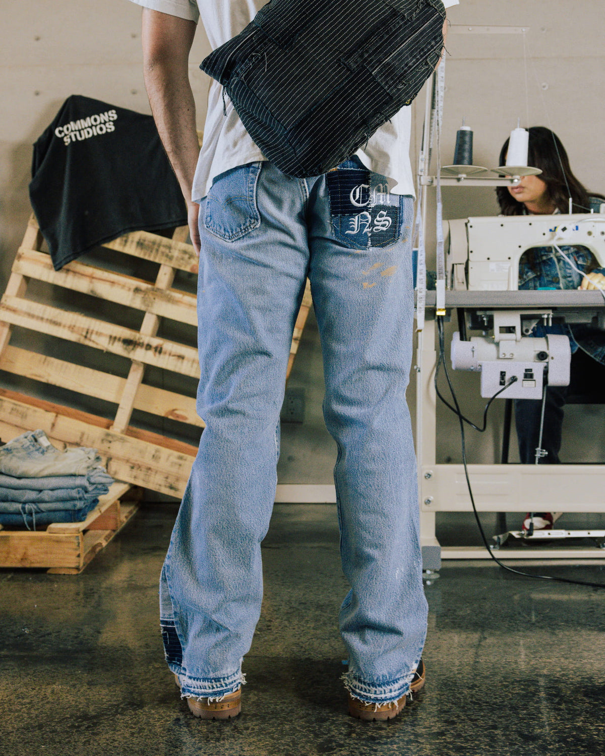 Person wearing light blue wide-leg jeans with patchwork pockets and frayed hems in a sewing workshop.