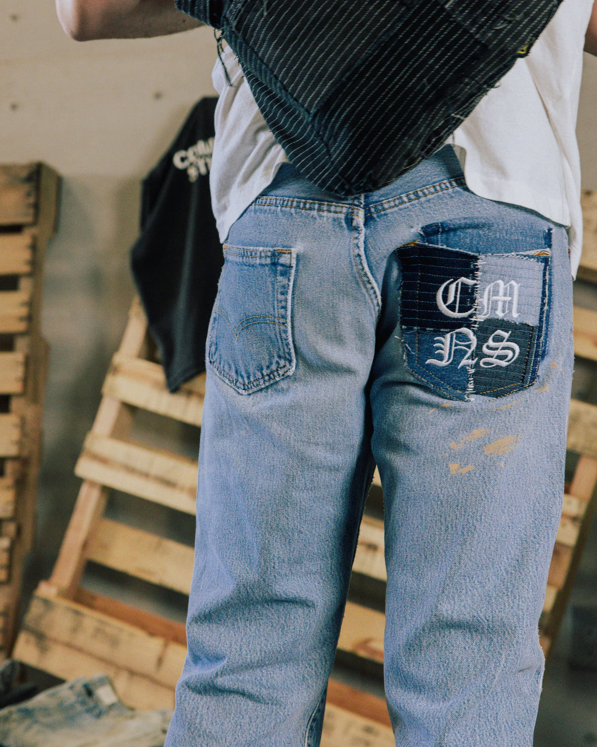 Person wearing light blue denim jeans with a patchwork back pocket and white embroidered lettering beside wooden pallets.
