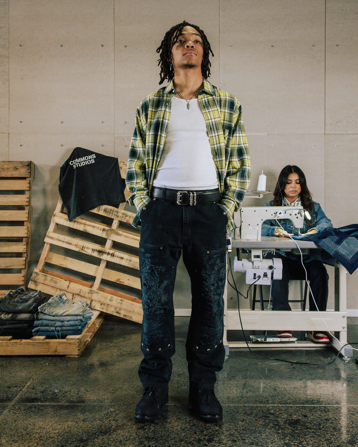 Model wears a green and yellow plaid shirt, white tank, and dark jeans with textured front panels in a sewing workshop.