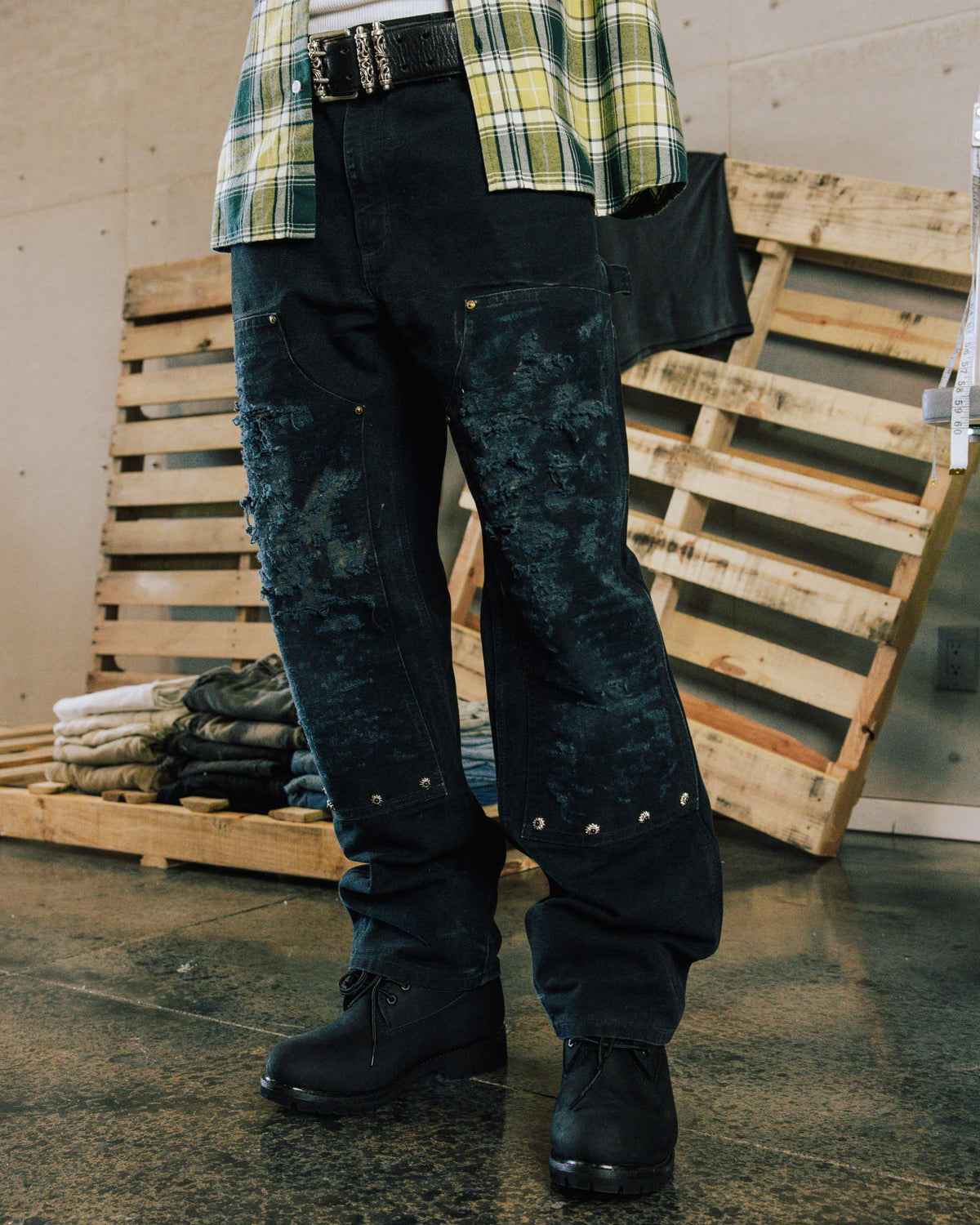 Model wears distressed dark denim jeans with studded panels, a plaid shirt and black boots beside wooden pallets.