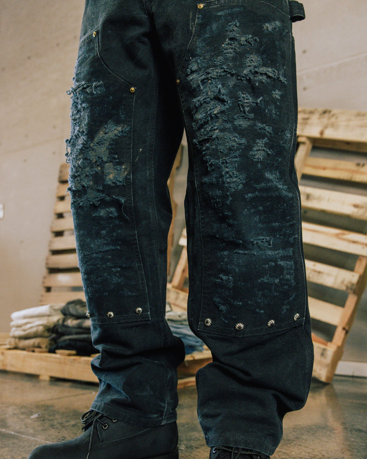 Dark blue distressed denim jeans with metal studs and black lace-up boots, shown beside wooden pallets.