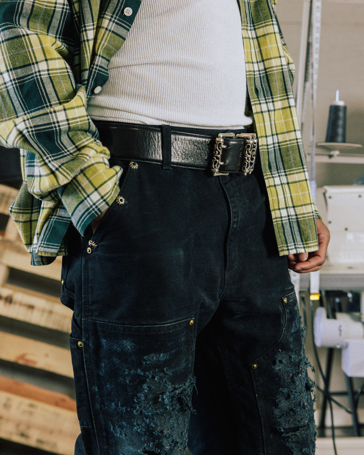 Person wearing distressed black denim cargo pants, an ornate black leather belt, and a green and yellow plaid shirt.