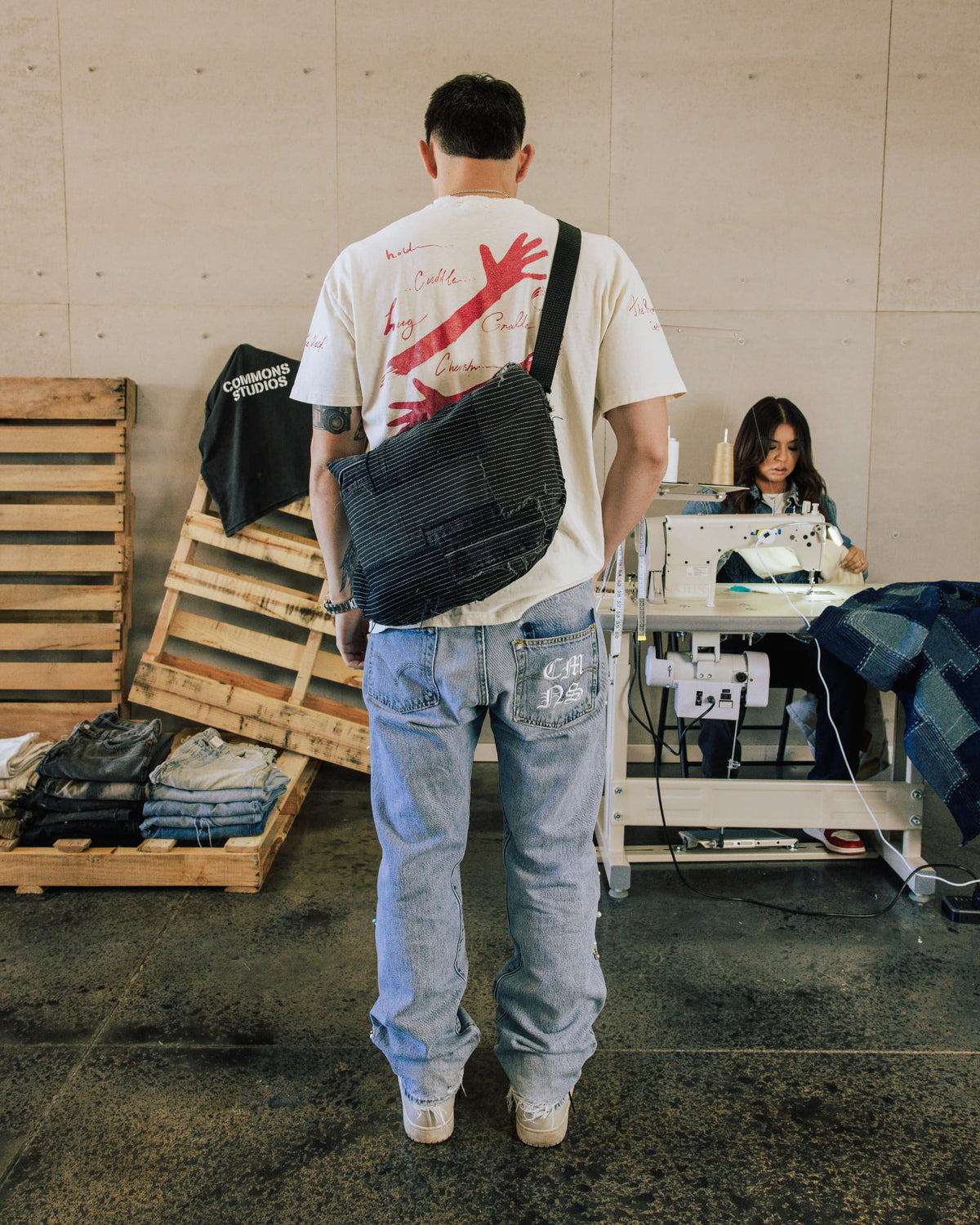 Man wearing a white graphic T-shirt, faded blue jeans and a dark striped crossbody bag in a sewing workshop.