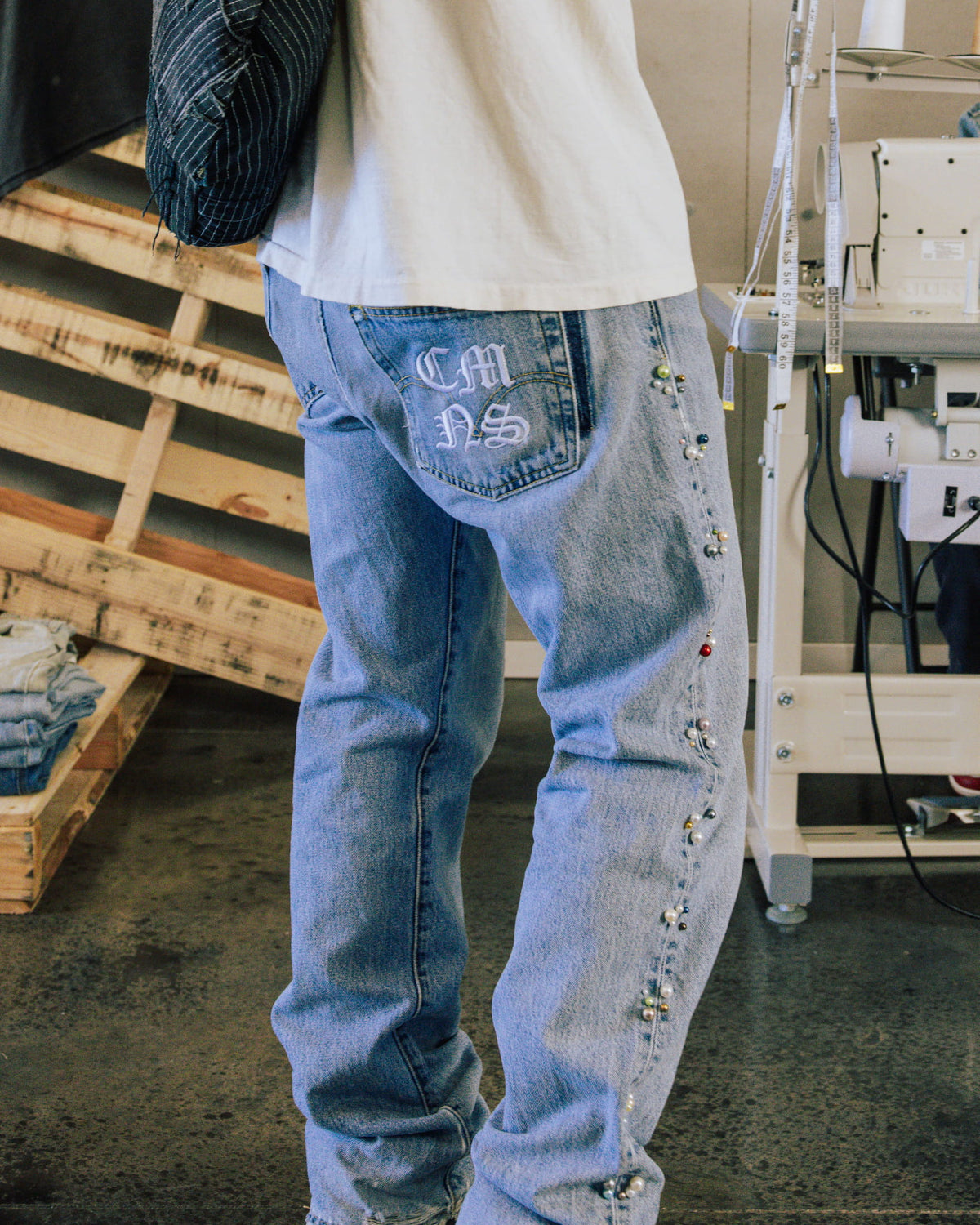 Light blue denim jeans with embroidered back pocket and beaded side seams, worn beside a sewing machine.