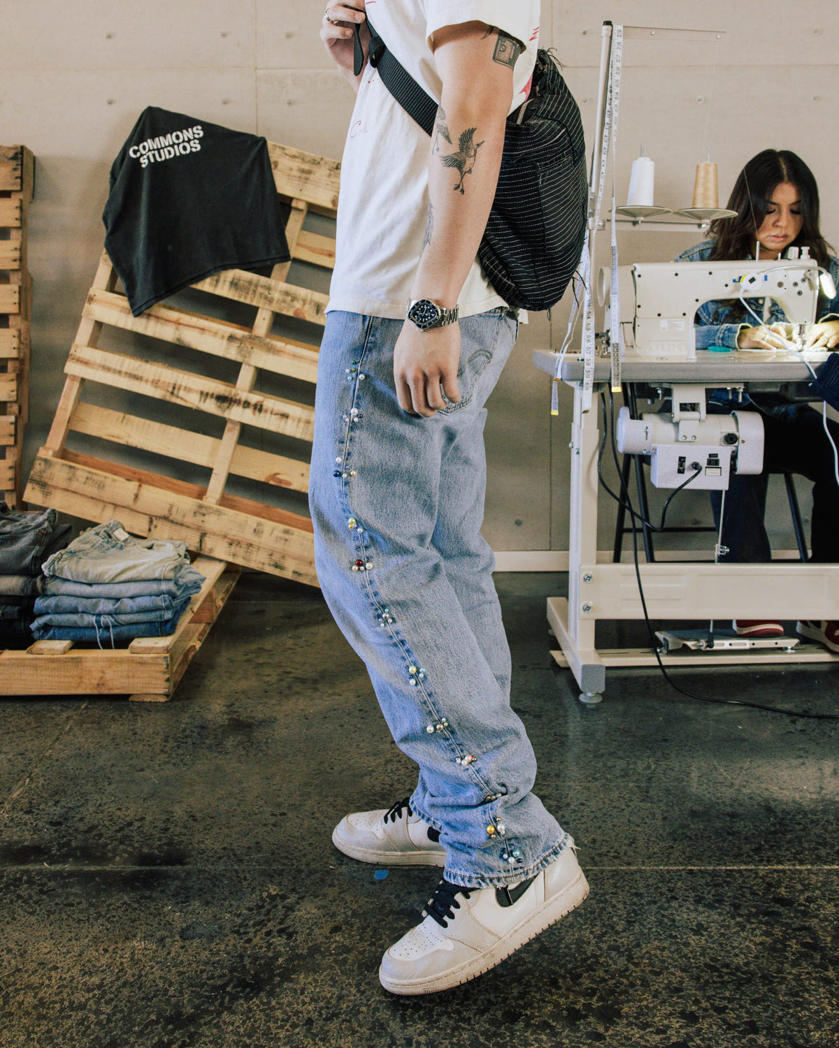 Person wearing light blue denim jeans with beaded side seams, a white T-shirt and black shoulder bag in a sewing studio.